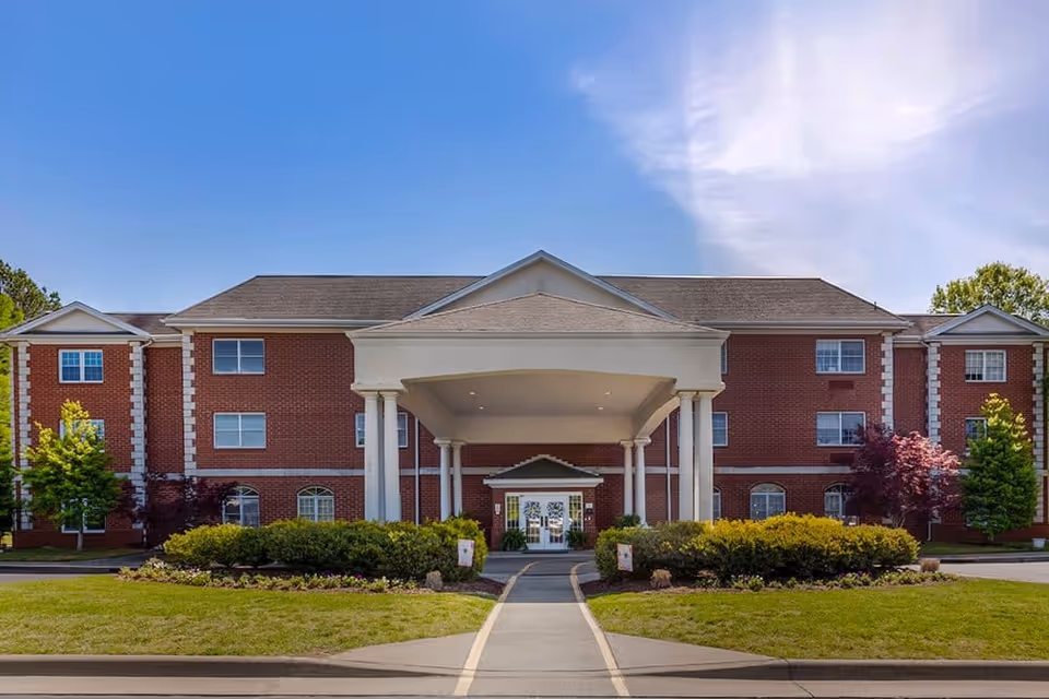 Front exterior view of a three-story brick building with a large covered entrance supported by white columns, surrounded by neatly trimmed bushes and trees under a clear blue sky.