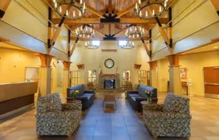 Spacious living room area with high wooden beam ceilings and multiple circular chandeliers. The room features two patterned armchairs and two dark sofas arranged around a central coffee table. A fireplace with a clock above it is centered on the far wall, flanked by built-in shelves. The room has warm yellow walls and tiled flooring.