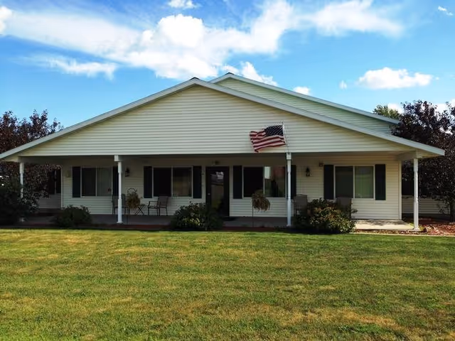 Front view of a single-story ranch-style building with a covered porch, an American flag, and a grassy lawn.