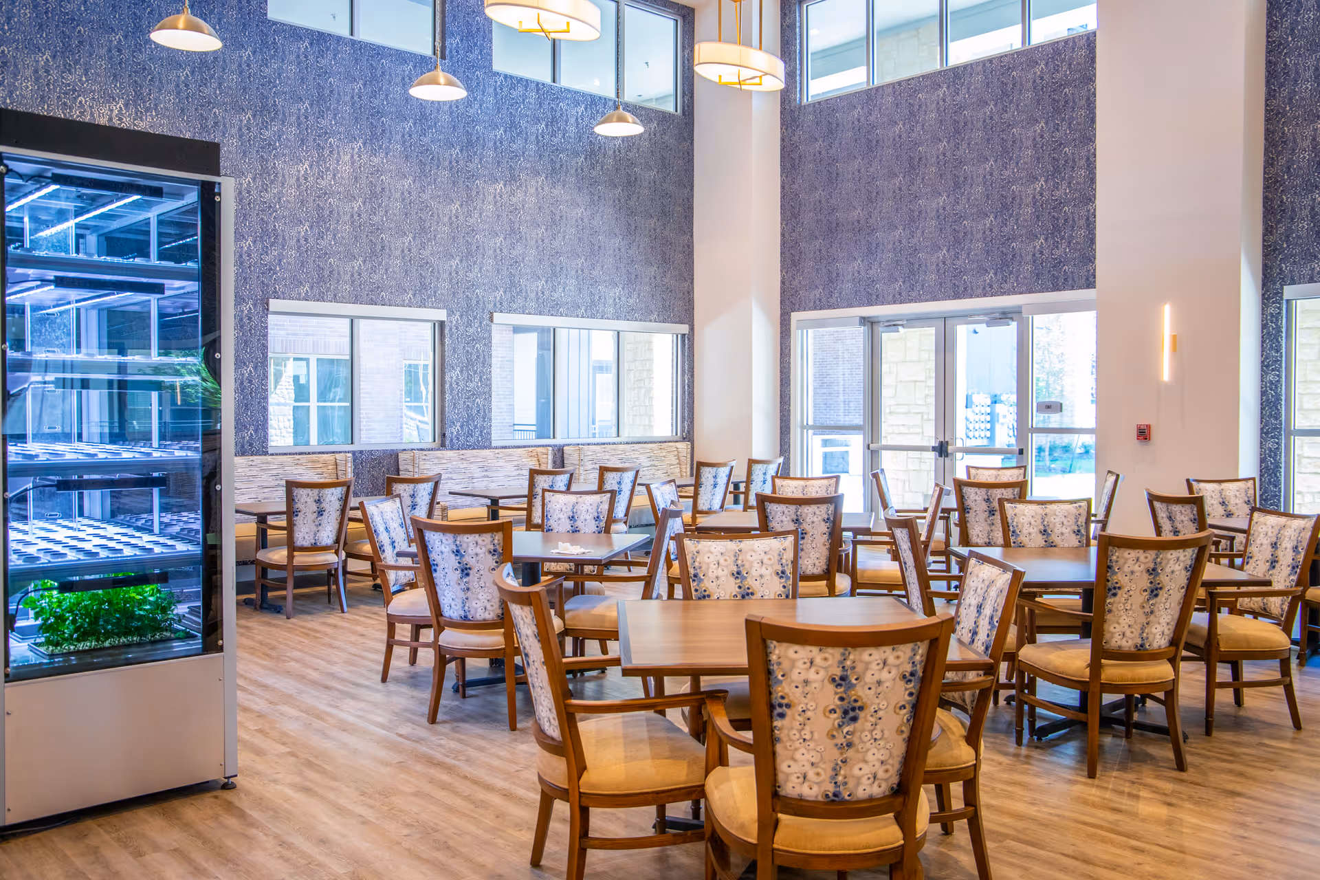 A bright dining room with multiple wooden tables and chairs featuring patterned upholstery. The room has large windows allowing natural light to fill the space, high ceilings, and modern pendant lights hanging from above. There is a vertical indoor garden or plant display on the left side of the image.