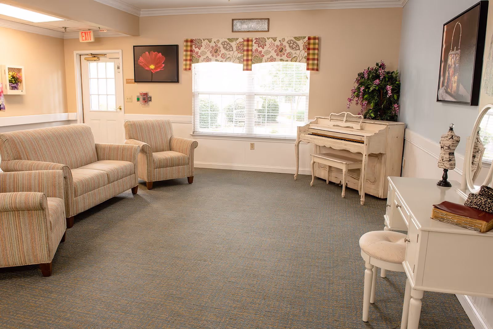 A cozy living room area in a senior living facility featuring a striped sofa and armchair, a white vintage piano with a matching bench, a white vanity table with a round mirror and cushioned stool, floral artwork on the walls, a window with floral and plaid valances, and a potted plant with purple flowers in the corner.
