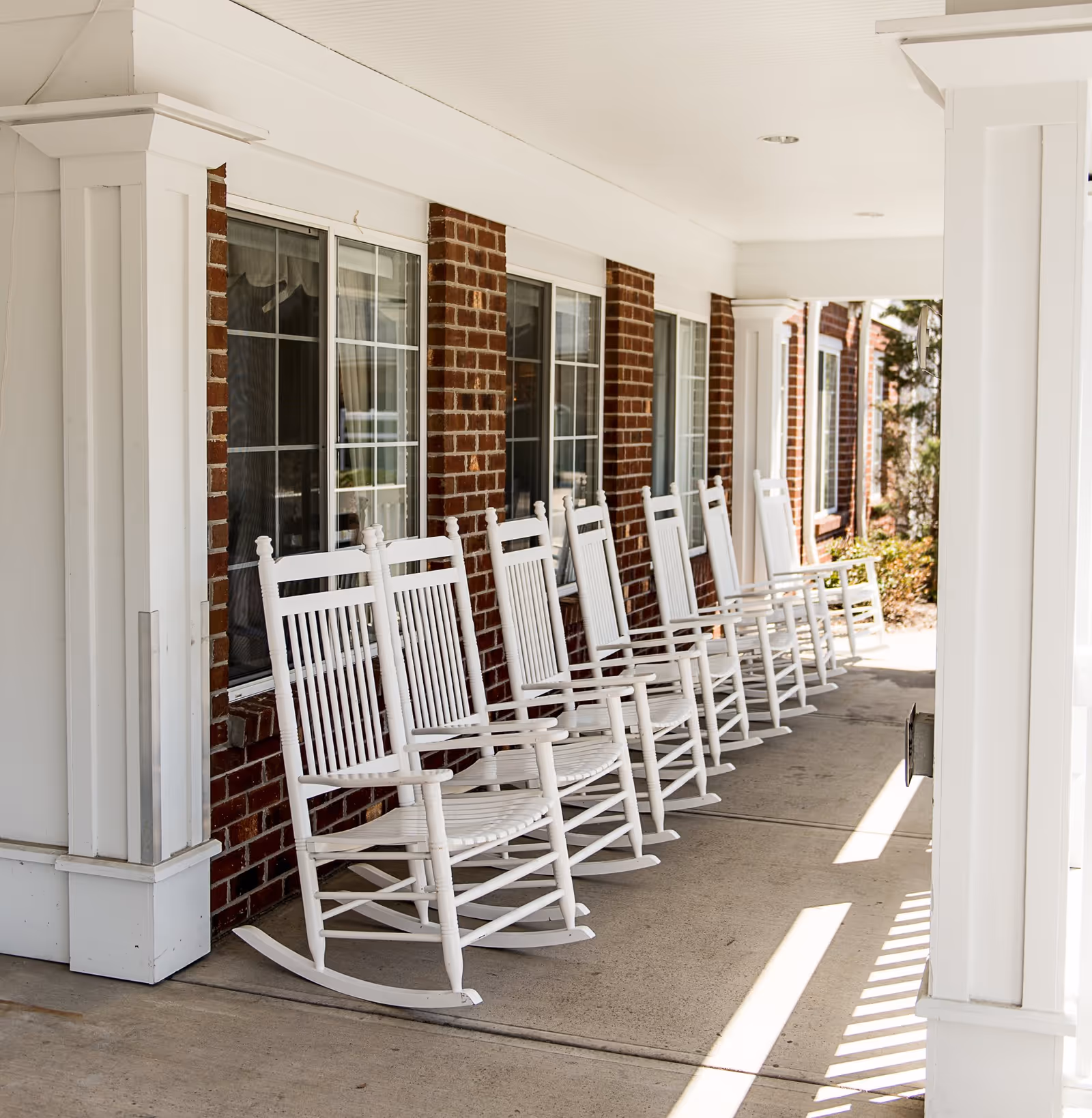 A row of white wooden rocking chairs lined up on a covered porch outside a brick building with white trim and multiple windows.