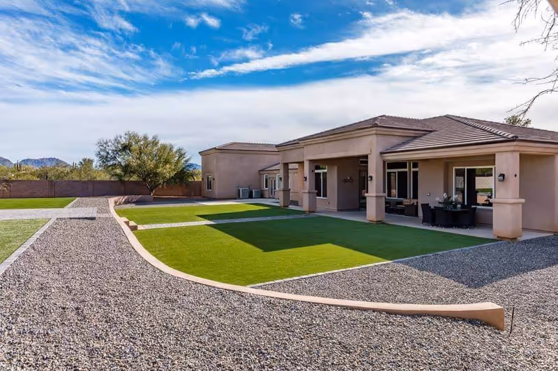 Exterior view of a single-story building with a covered patio area, green lawn, gravel pathways, and desert landscaping under a partly cloudy blue sky.