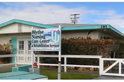 Exterior view of Blythe Nursing Care Center & Rehabilitation Services building with a white fence and a sign displaying the facility name and services. The building has a light green roof and beige walls, with some bushes and a clear blue sky in the background.