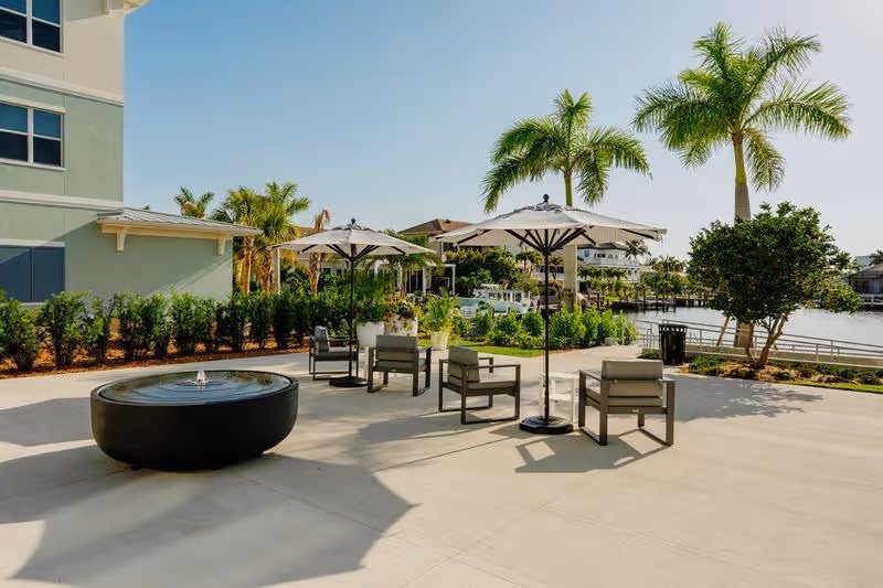 Outdoor patio with seating, umbrellas, a round fountain, palm trees, and a waterfront view.