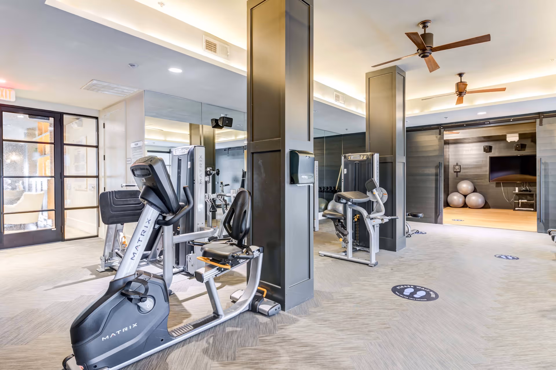 A modern fitness room with exercise equipment including a recumbent bike and weight machines. The room has large mirrors on the walls, ceiling fans, and a separate area with exercise balls and a TV. The floor has social distancing markers.