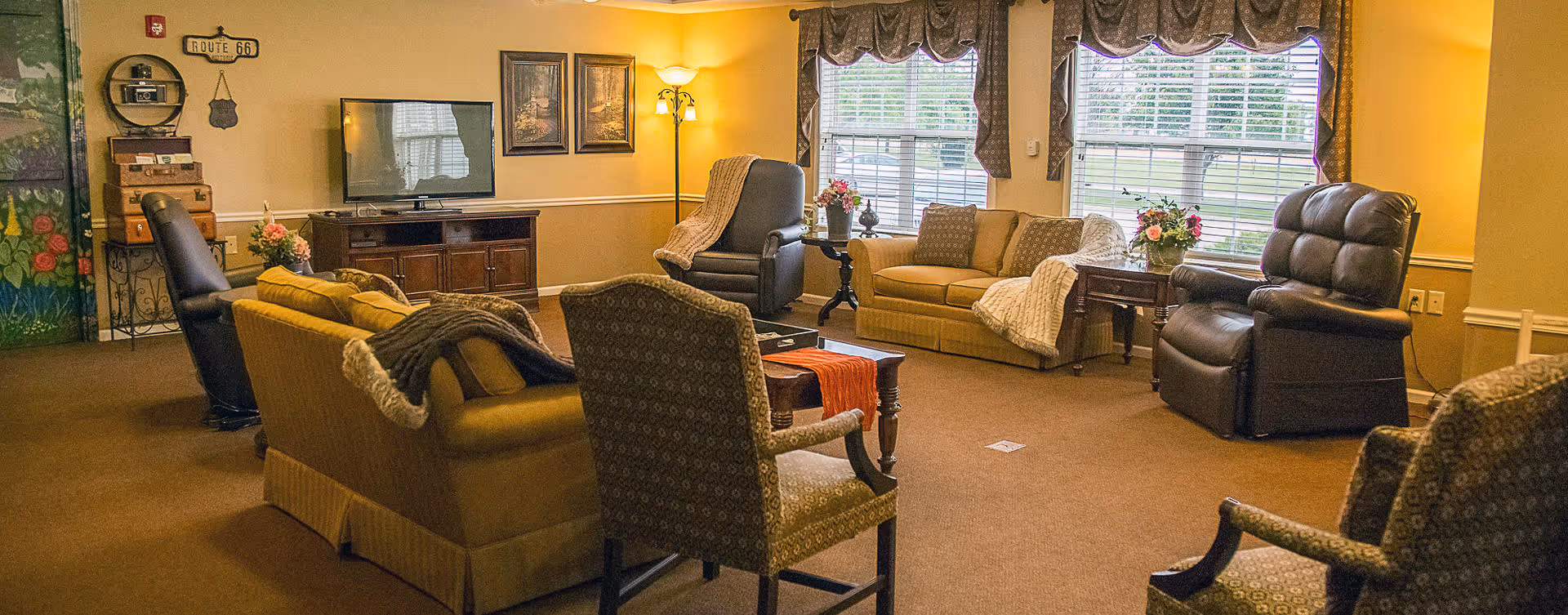 A cozy living room area with multiple armchairs and sofas arranged around a central coffee table. The room has warm yellow walls, large windows with valances letting in natural light, decorative flowers on side tables, a flat-screen TV on a wooden cabinet, and framed artwork on the walls.