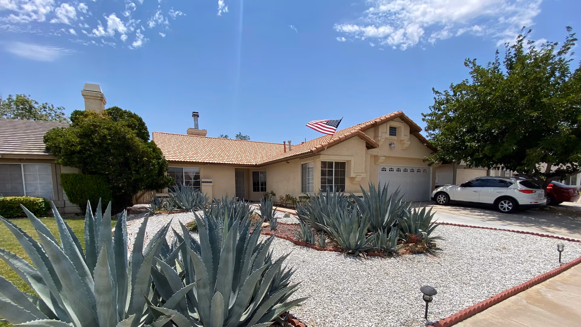 Single-story residential building with a beige exterior and a tiled roof, featuring an American flag on a pole in the front yard. The yard is landscaped with large agave plants and white gravel, with a driveway on the right side where two cars are parked. Trees and bushes surround the house under a partly cloudy blue sky.