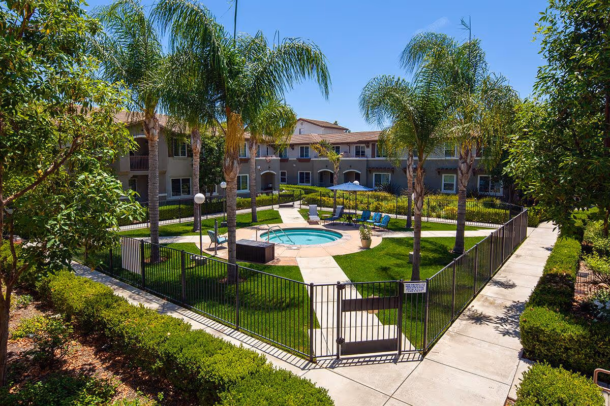Outdoor courtyard area at Woodland Village featuring a fenced circular hot tub surrounded by palm trees, lounge chairs, and a patio umbrella, with a two-story residential building in the background under a clear blue sky.