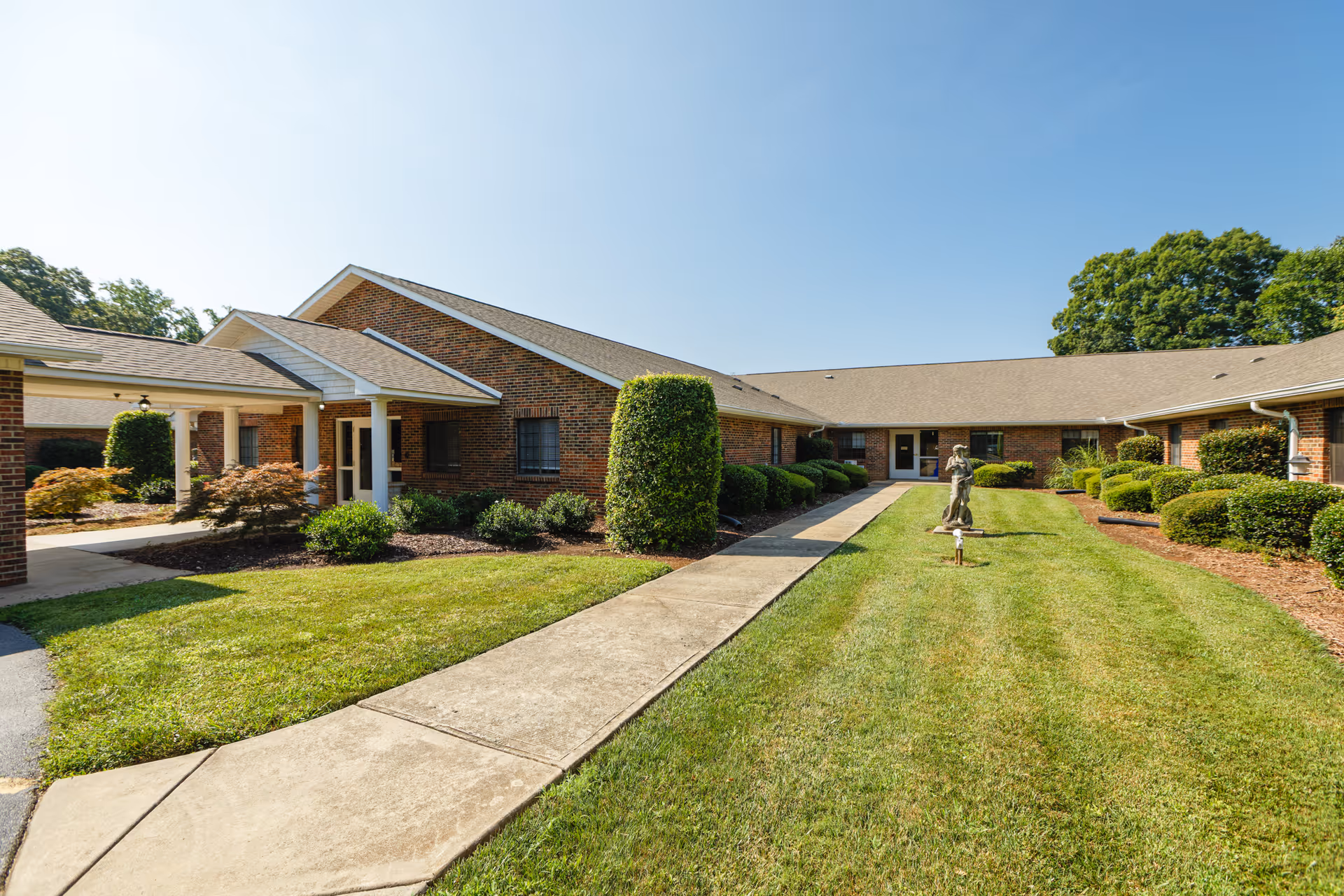 A sunny outdoor courtyard area of a senior living facility with a concrete walkway running through the middle, surrounded by well-maintained green grass and neatly trimmed bushes. The building is a single-story brick structure with multiple doors and windows facing the courtyard. A small statue is placed on the grass near the walkway.