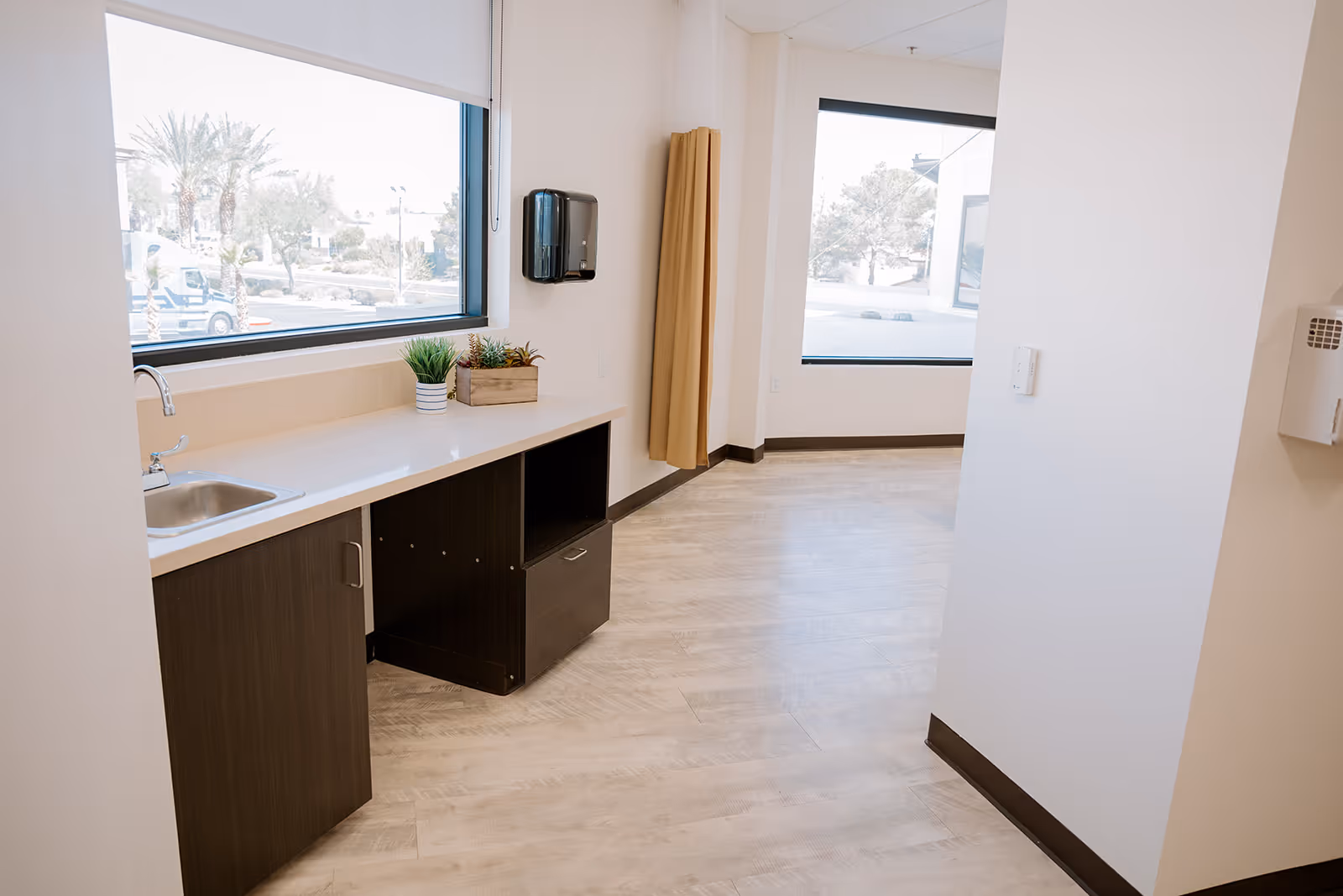 A clean and bright interior space with light wood flooring, a countertop with a small sink, two potted plants, and large windows letting in natural light. The walls are white, and there is a beige curtain hanging near one window.