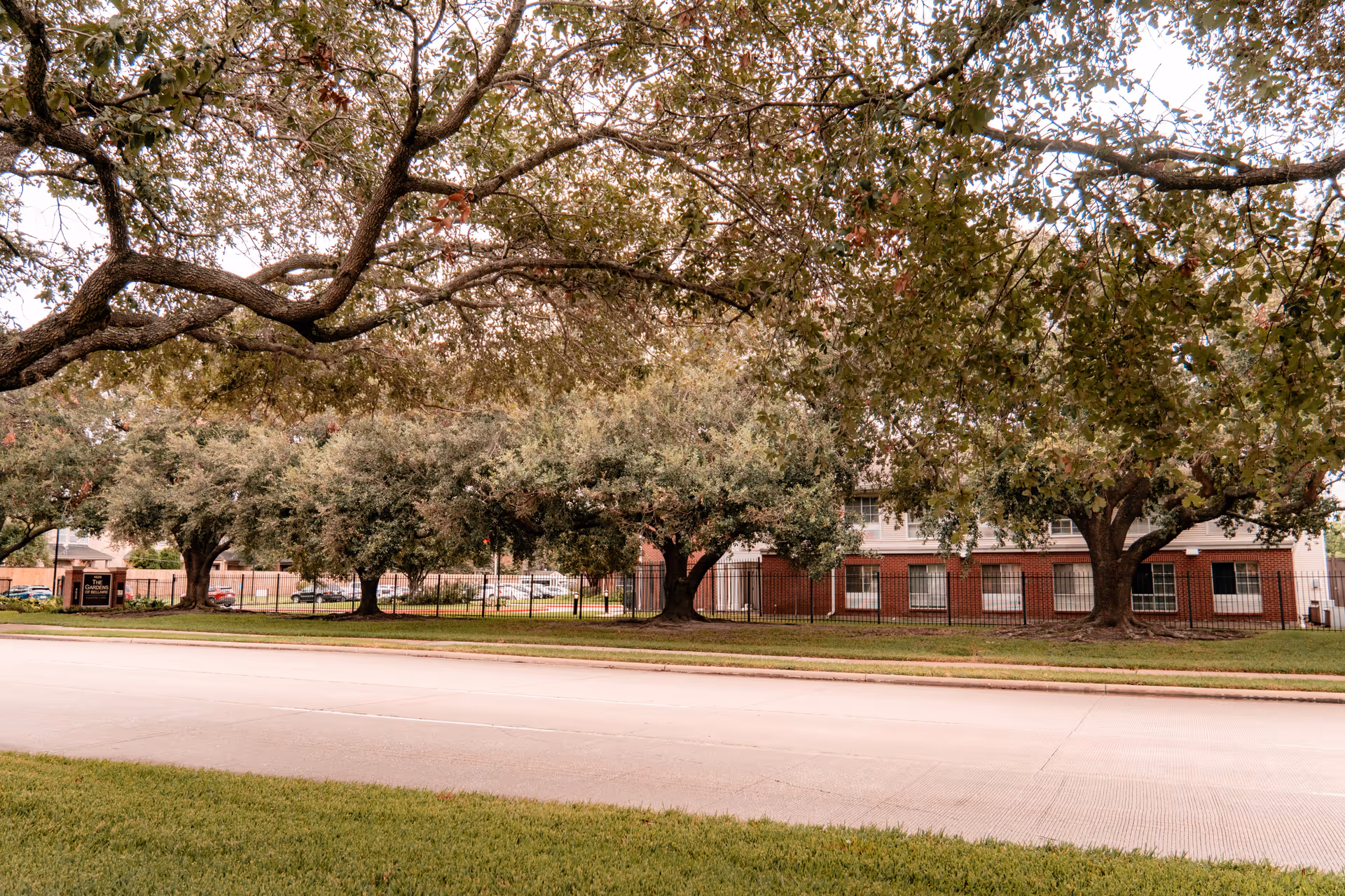 View of a senior living facility named The Gardens of Bellaire, showing a street in the foreground, a grassy area, several large trees, and a two-story brick and white building behind a black metal fence.