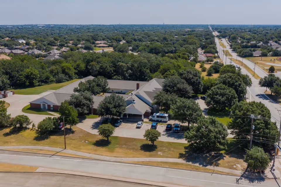 Aerial view of Deer Creek Senior Living facility surrounded by trees and greenery, with a parking lot and several vehicles in front. The building is a single-story structure with a gray roof, situated near a road with other residential houses in the background.