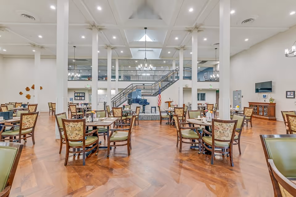 Spacious dining room with multiple tables and chairs arranged neatly on a polished wooden floor. The room features high ceilings with recessed lighting and chandeliers, white pillars, a staircase leading to an upper level, a grand piano, and an American flag near a small stage area.