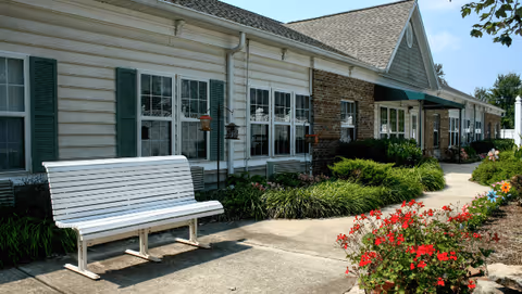 Exterior view of a senior living facility building with white siding and brick accents, featuring multiple windows with green shutters. A white bench is placed on a concrete walkway surrounded by green shrubs and red flowers under a clear blue sky.