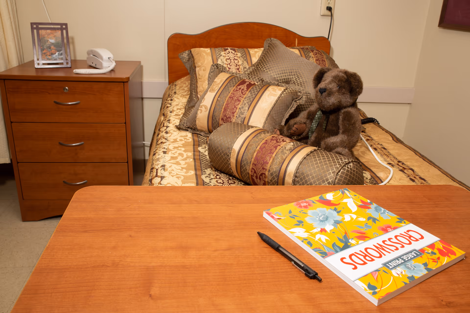 A cozy bedroom with a single bed adorned with multiple decorative pillows and a teddy bear sitting on it. Next to the bed is a wooden nightstand with a white telephone and a framed picture. In the foreground, there is a wooden table with a colorful crossword puzzle book and a black pen on it.