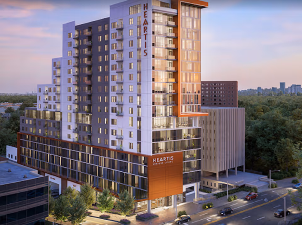 Exterior view of a modern multi-story building named Heartis Buckhead Senior Living during dusk, surrounded by trees and nearby buildings, with cars on the street in front.