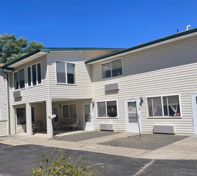 Exterior view of a two-story beige siding building with multiple windows and doors, a covered porch area with chairs, and a paved driveway in front under a clear blue sky.
