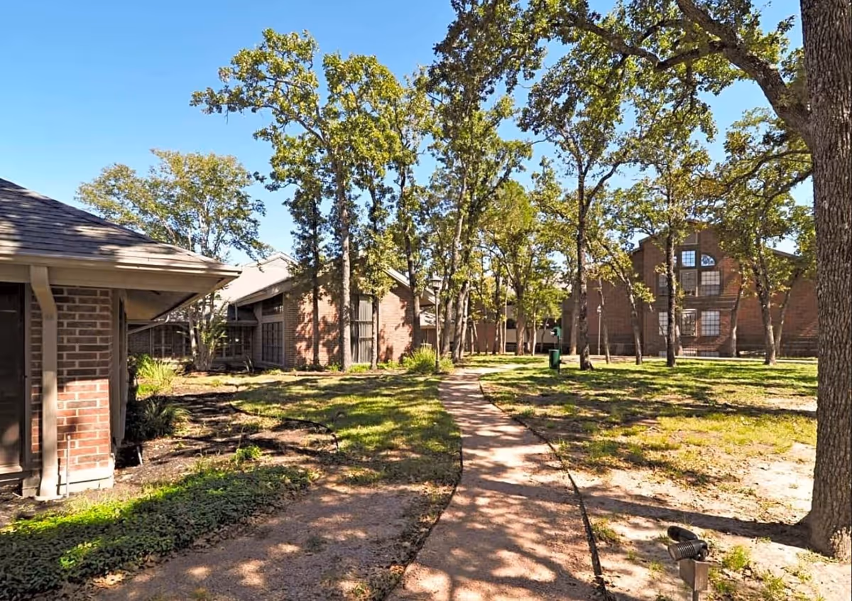 A sunny outdoor area at Serenity at Briarcrest featuring a paved walkway winding through a grassy yard with several mature trees. Surrounding the yard are multiple brick buildings with large windows, under a clear blue sky.