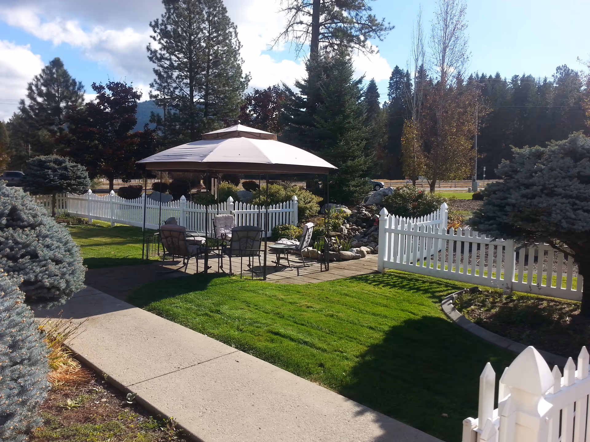 Outdoor seating area with a large umbrella covering a table and several chairs, surrounded by a white picket fence, green grass, trees, and shrubs under a partly cloudy sky.