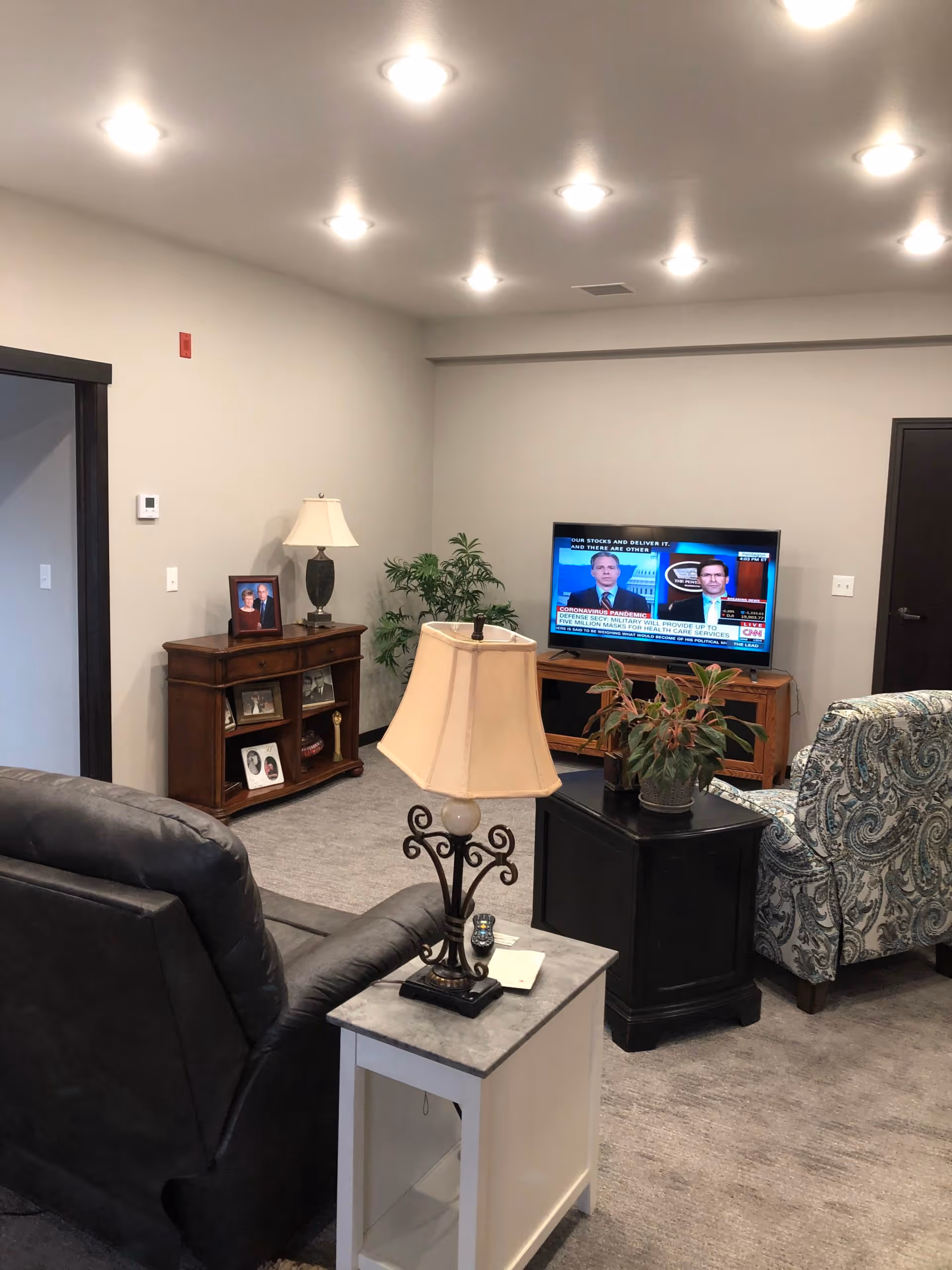 A cozy living room with a gray recliner chair and a patterned armchair facing a flat-screen TV mounted on a wooden stand. There is a small white side table with a decorative lamp and a remote control next to the recliner. A wooden cabinet with framed photos and a lamp is against the wall, and a green potted plant adds a touch of nature to the room. The walls are painted light gray and the floor is carpeted.