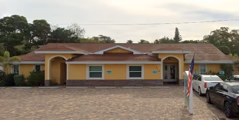 Single-story yellow guest home with arched entrances, brick trim, a paved parking area with cars, and an American flag near the front.
