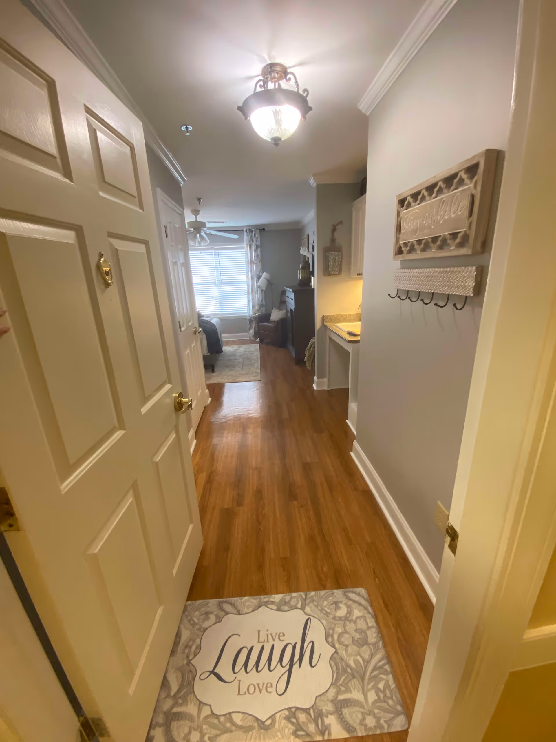 View through an open door into a hallway with wooden flooring leading to a bedroom area with a window covered by blinds and curtains. A decorative mat on the floor near the door reads 'Live Laugh Love'. On the right wall, there is a decorative wall hanging with hooks. The hallway also features a small countertop with cabinets above it and a ceiling light fixture.