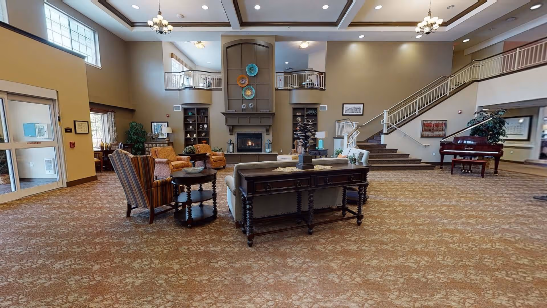 Spacious two-story senior living facility lobby with seating areas, a central fireplace, staircase and a grand piano.