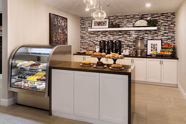 A modern cafe-style area with a display case filled with desserts and pastries on the left, a central counter with plates of assorted pastries, and a back counter with coffee dispensers, a water dispenser, fruit, and decorative items against a tiled backsplash wall.