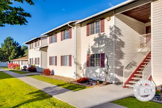 Exterior view of a two-story apartment building with beige siding and red shutters. There is a concrete walkway and green lawn in front, with some small bushes and trees. A staircase with red steps leads to the second floor on the right side of the building. The sky is clear and blue.