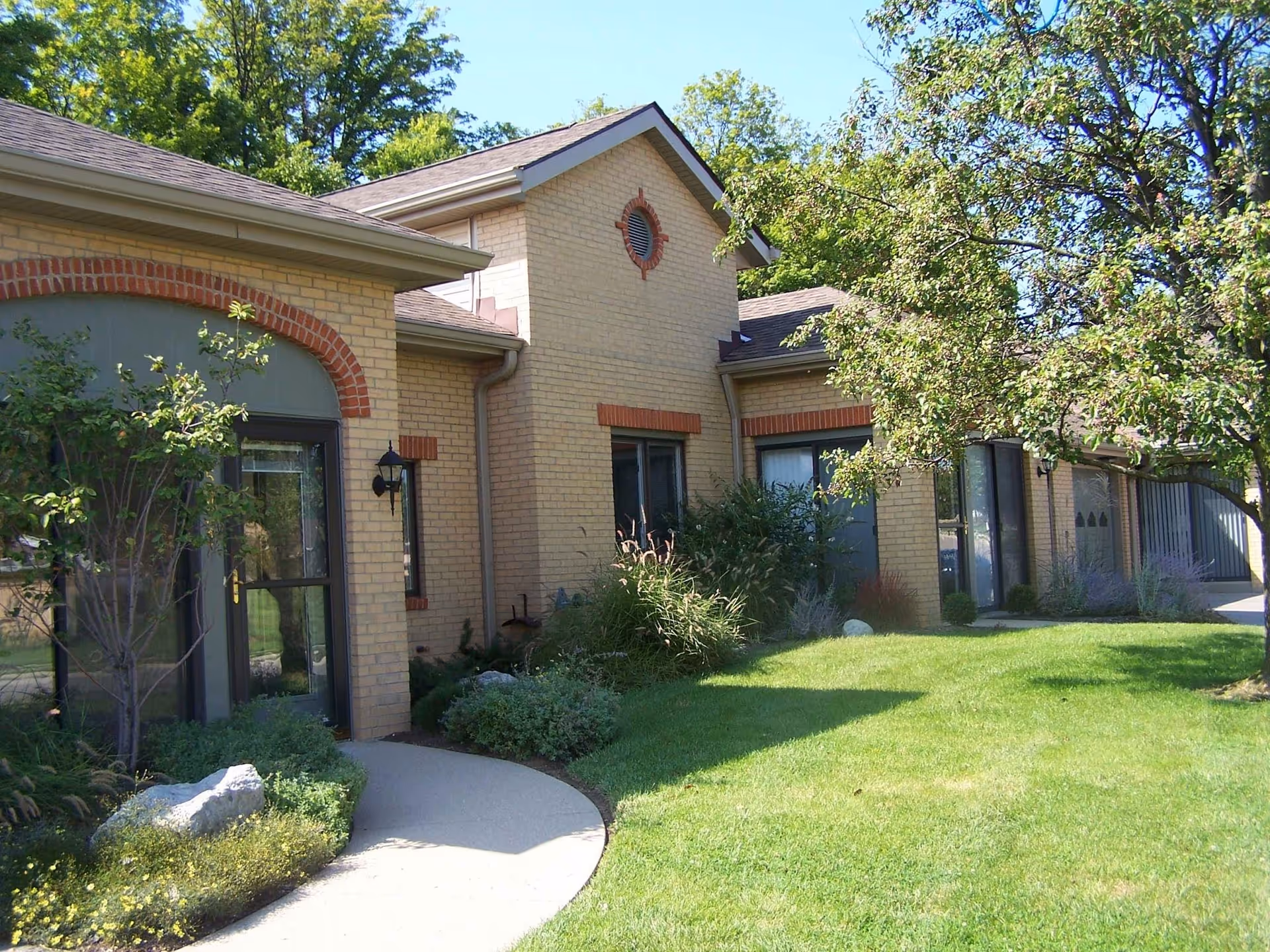 Brick one-story building with arched windows, landscaping, and a curved walkway across a green lawn under trees.
