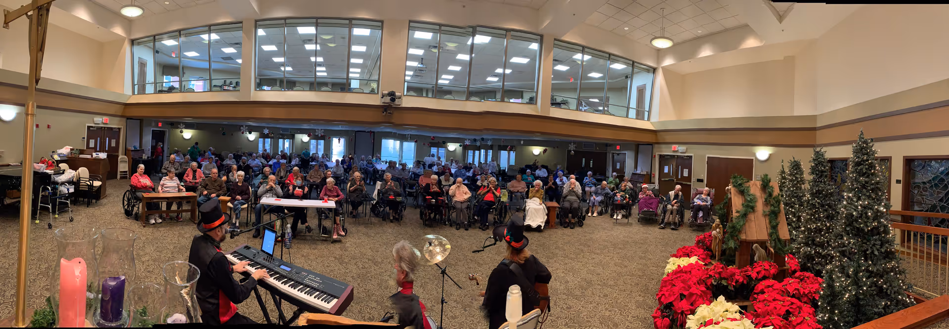 A large group of elderly residents seated in a spacious, well-lit common area of a senior care facility, watching a live musical performance by three musicians, one playing a keyboard and another playing a guitar. The room is decorated with Christmas trees, poinsettias, and holiday greenery, creating a festive atmosphere.