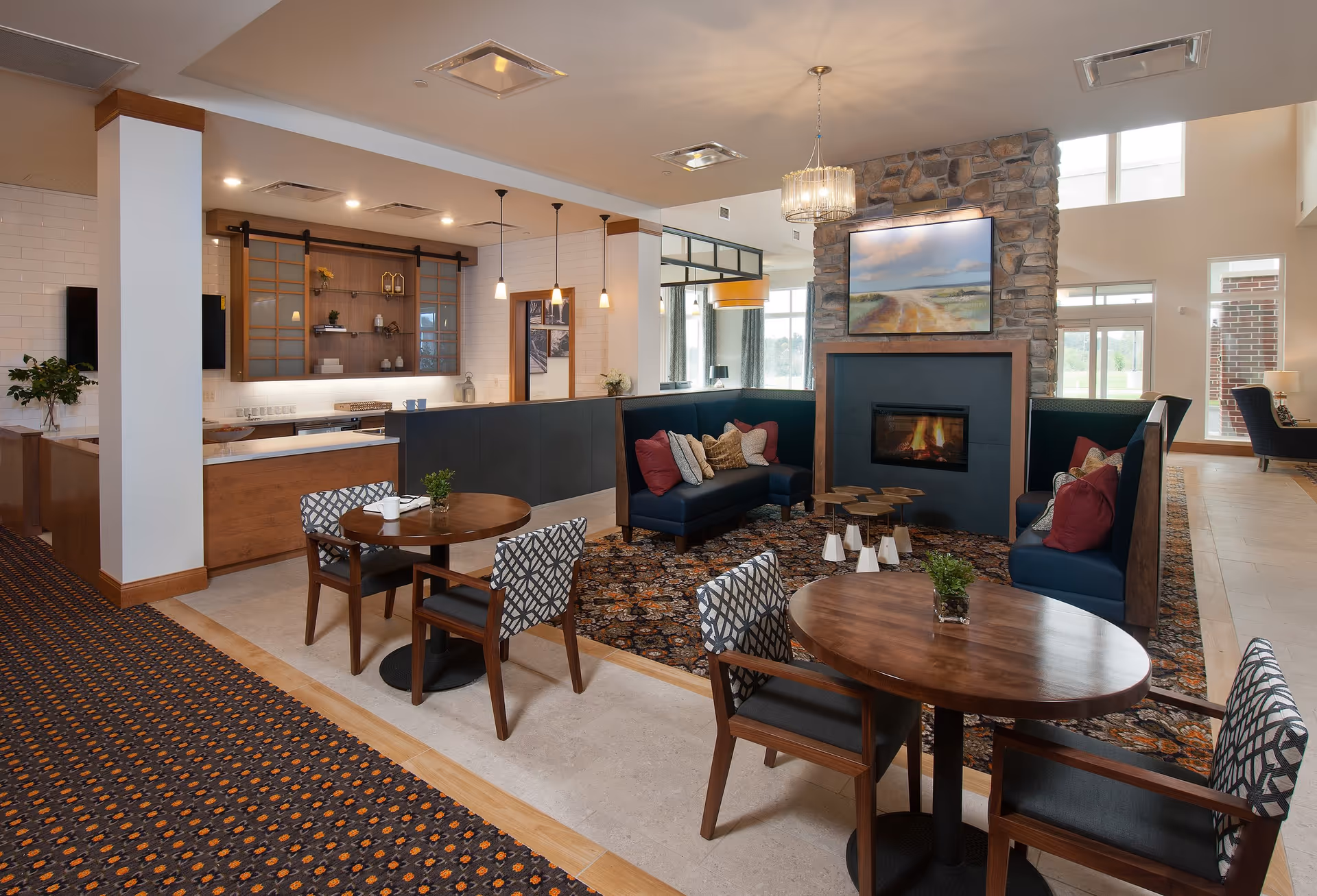 A cozy common area in a senior living facility featuring a stone fireplace with a painting above it, surrounded by blue cushioned seating with red and beige pillows. There are round wooden tables with patterned chairs, a patterned carpet, and a kitchen area with pendant lights and wooden cabinetry in the background. Large windows allow natural light to fill the space.