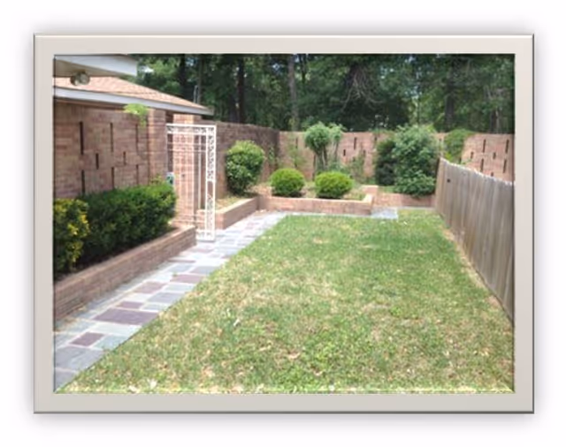 A small, enclosed outdoor garden area with a green lawn, bordered by a stone pathway on the left and a wooden fence on the right. There are brick walls surrounding the garden with various green shrubs and small trees planted along the edges. A white metal gate is visible on the left side near the brick wall.