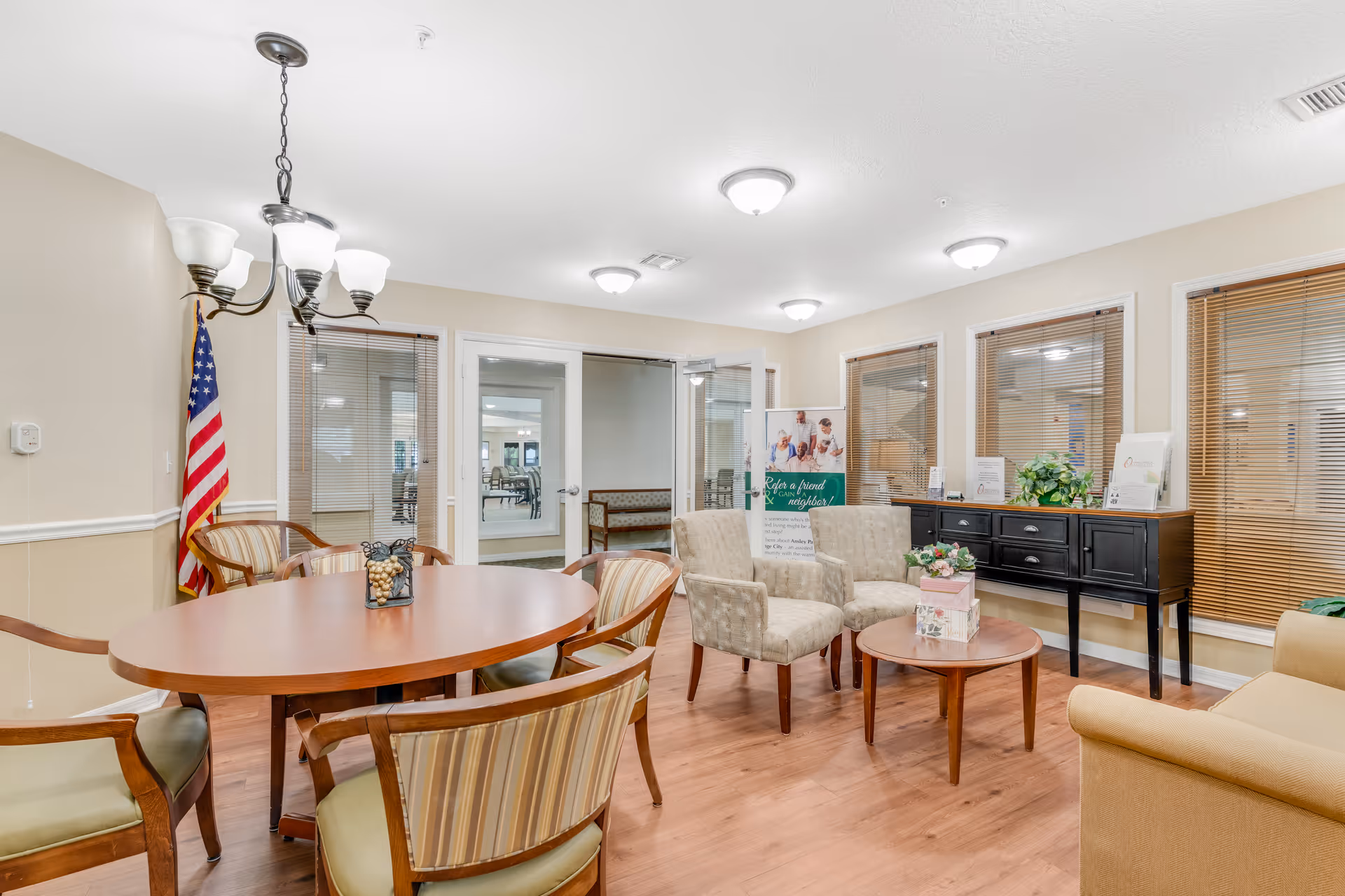 A well-lit common area in a senior living facility featuring a round wooden table with four striped cushioned chairs, two beige upholstered armchairs, a small round wooden side table with decorative items, a black sideboard with plants and brochures, and an American flag in the corner. The room has wooden flooring, beige walls, multiple windows with blinds, and glass doors leading to another room.
