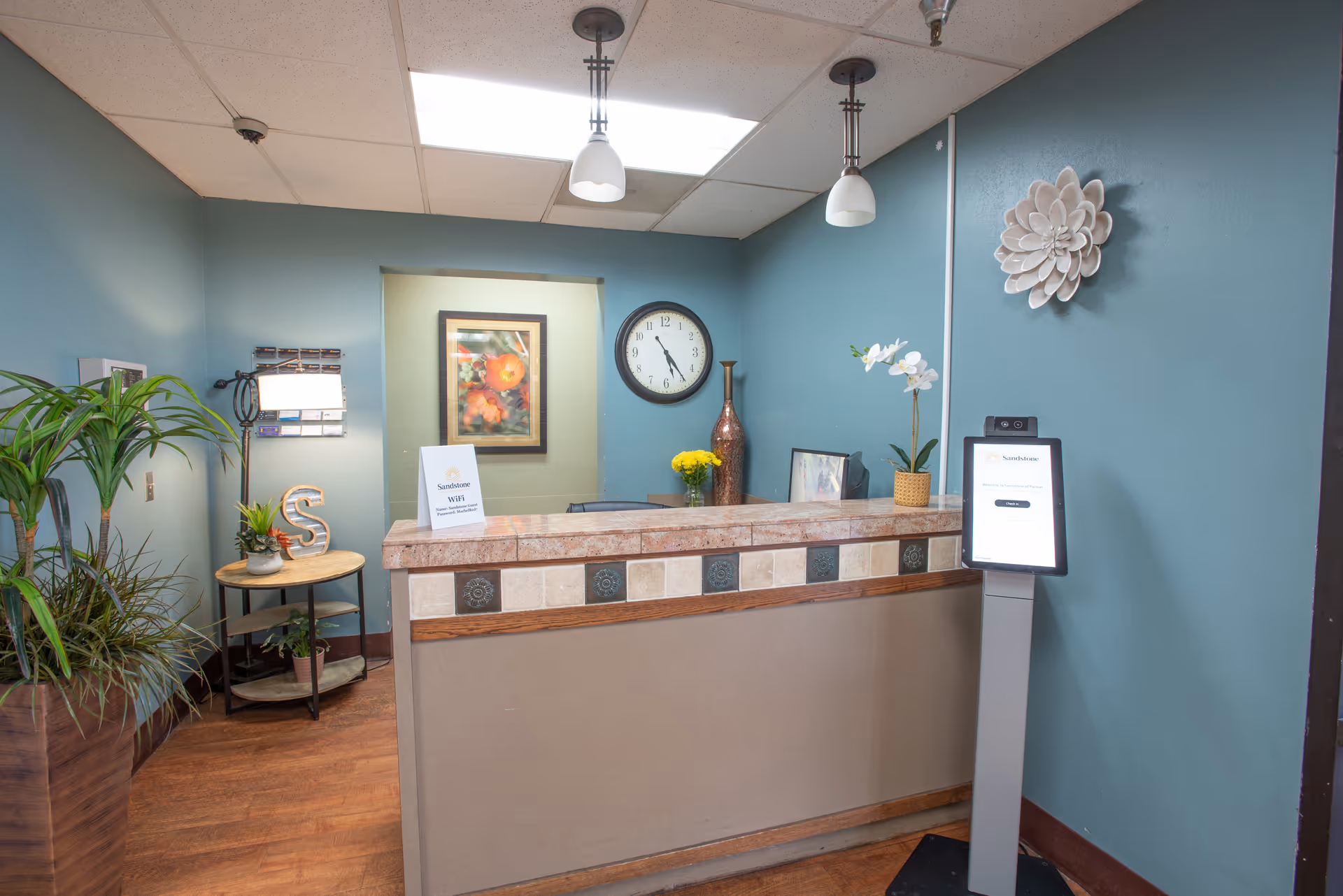Reception area with a tiled counter, a clock on the wall showing 11:05, decorative plants, a framed picture, and a digital check-in kiosk. The walls are painted teal and there are two hanging pendant lights above the counter.