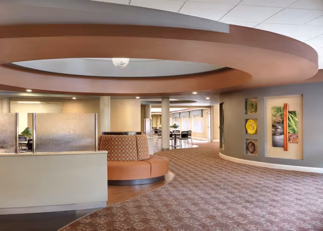 Interior view of a senior living facility with a curved ceiling design and a circular seating area upholstered in brown and patterned fabric. The space features carpeted flooring with a floral pattern, a partial divider with frosted glass panels, and wall art including nature-themed pictures. In the background, there are tables and chairs near large windows allowing natural light to enter.