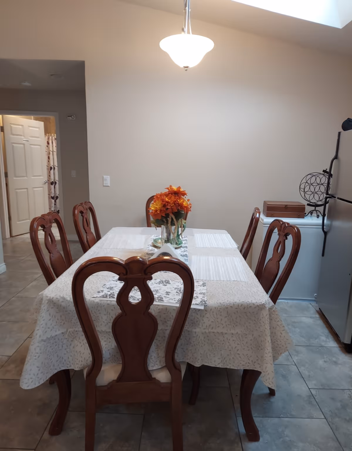 A dining room with a rectangular table covered with a light-colored tablecloth and six wooden chairs around it. A vase with orange and yellow flowers is placed in the center of the table. The room has tiled flooring, beige walls, and a hanging ceiling light. A refrigerator and a small cabinet are visible on the right side, and an open door leading to another room is on the left.