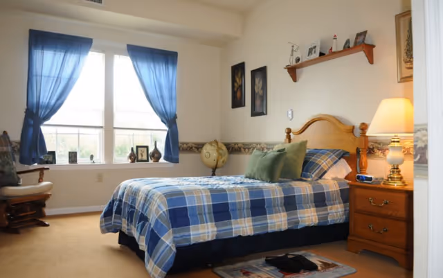 A cozy bedroom with a single bed covered in a blue and beige plaid comforter, two green pillows, and a wooden headboard. Next to the bed is a wooden nightstand with a lamp and an alarm clock. A small rug is placed on the floor near the bed. The room has beige walls with a decorative border, two framed pictures on the wall, a wooden shelf with small decorative items, and a globe on a stand. A window with blue curtains lets in natural light, and a cushioned rocking chair is positioned near the window.
