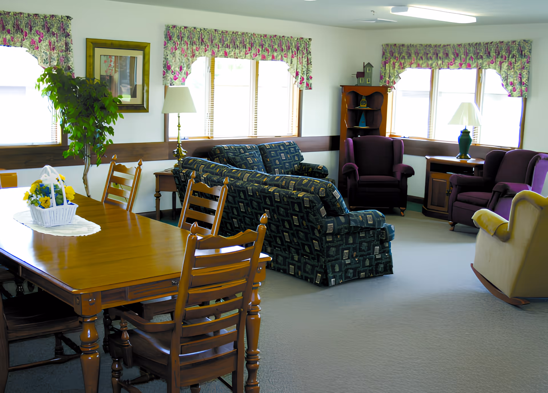 A bright living room area with multiple windows covered by floral valances. The room features a wooden dining table with chairs, a patterned sofa set, two purple armchairs, a beige rocking chair, a floor lamp, a table lamp, a small wooden shelf with decorative items, and a potted plant near the window.