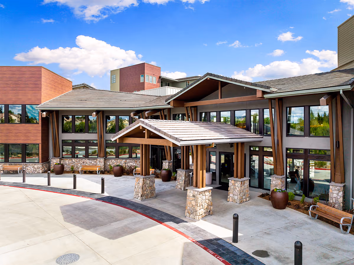 Front entrance of a modern building featuring a covered porte-cochere with stone pillars, large windows, and potted plants under a blue sky.