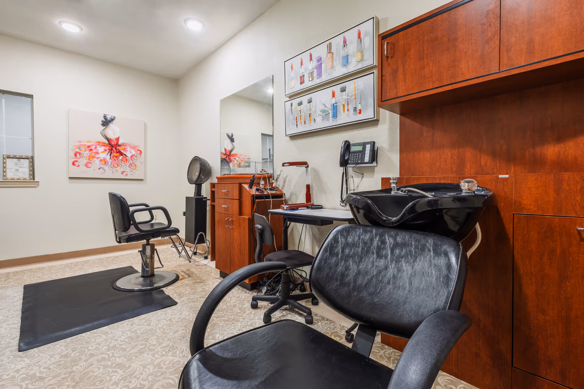 Interior view of a salon area in a senior living facility featuring black salon chairs, a hair washing station with a black sink, wooden cabinets, a large mirror, and colorful artwork on the walls. The room has beige patterned carpet and bright ceiling lights.