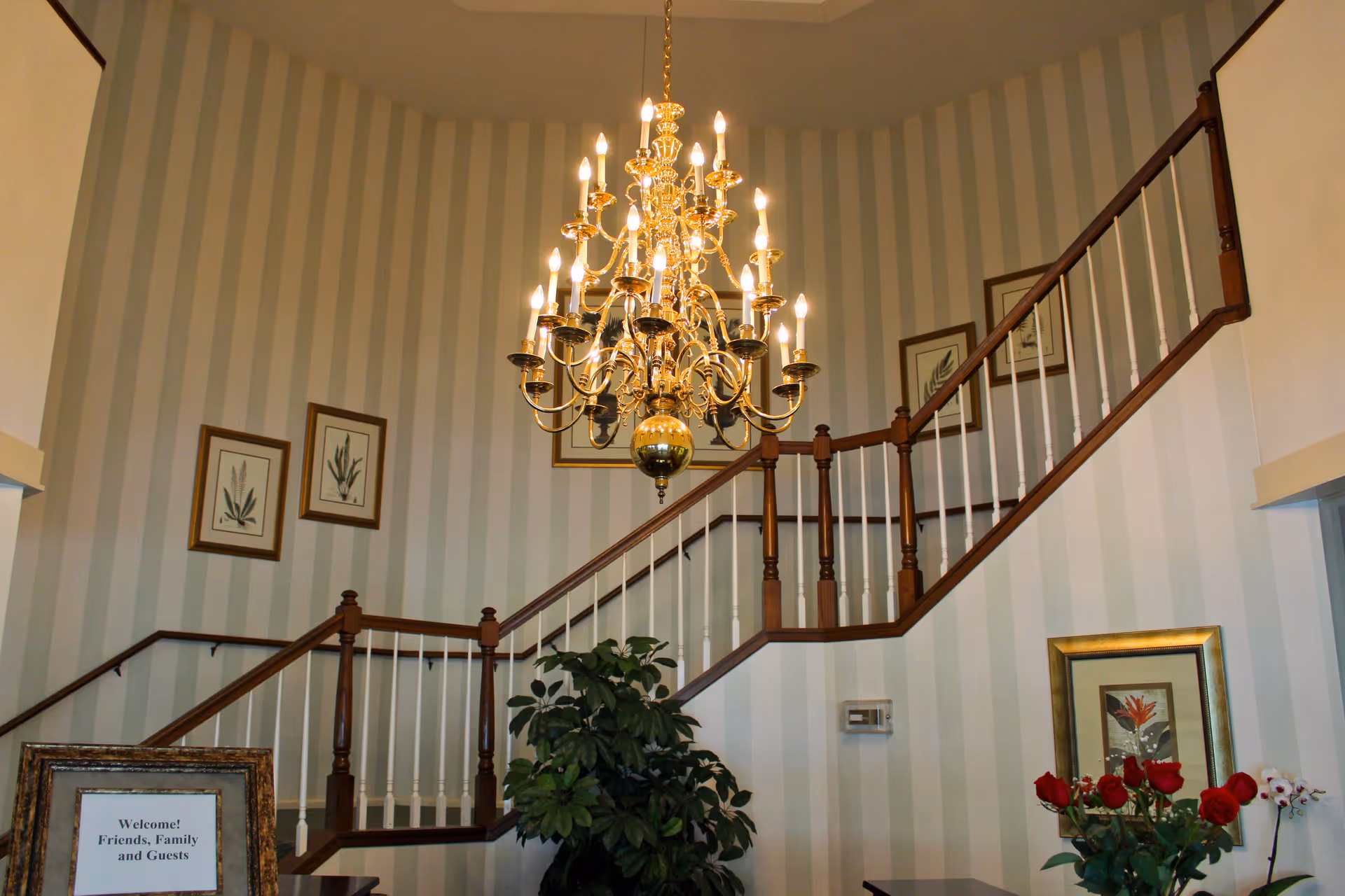 Interior view of a stairway with wooden handrails and white balusters, a large ornate chandelier hanging from the ceiling, striped wallpaper, framed botanical prints on the walls, a potted plant, and a table with a vase of red roses and a framed welcome sign.