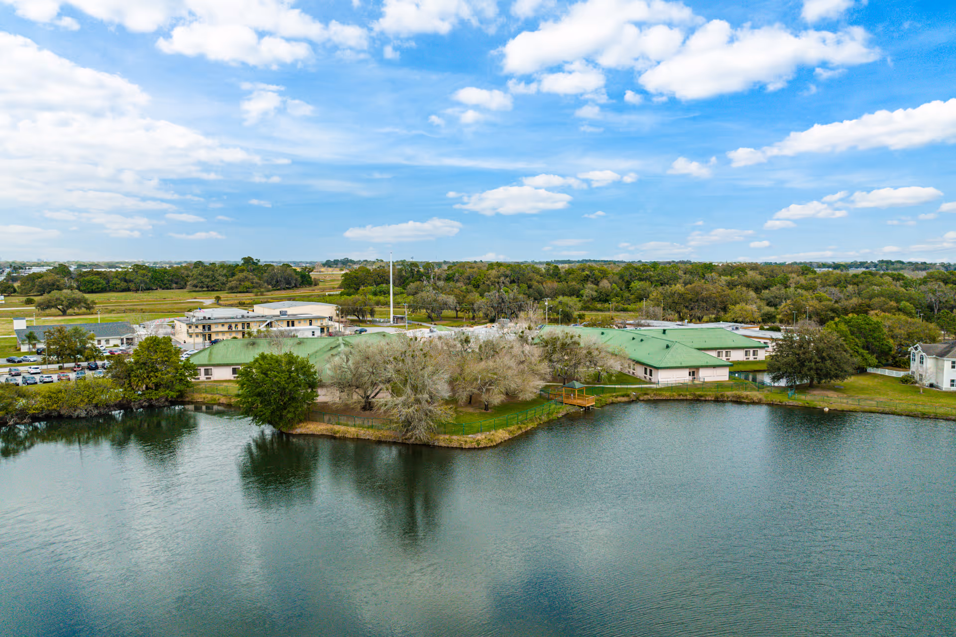 Aerial view of green-roofed senior living buildings on the shore of a lake with trees and a small dock.