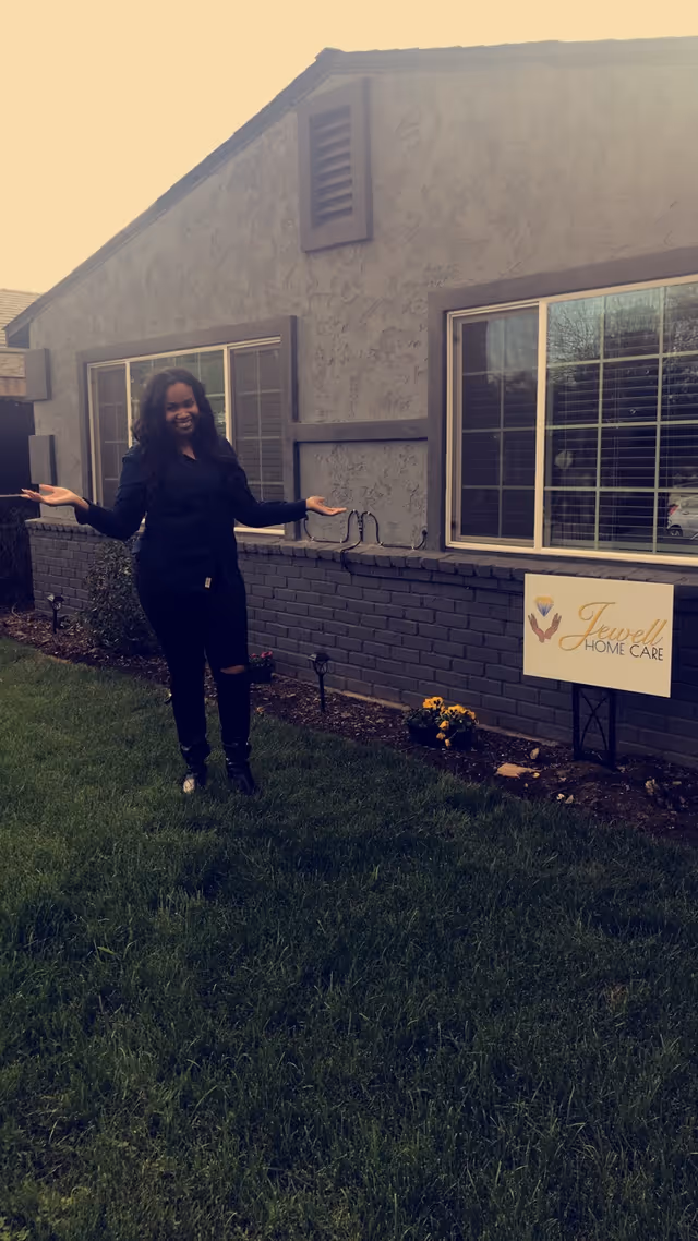A woman dressed in black stands on a green lawn in front of a gray brick and stucco building with two windows. She is smiling and gesturing with her hands towards a sign that reads 'Jewell Home Care' with a logo featuring hands and a heart.
