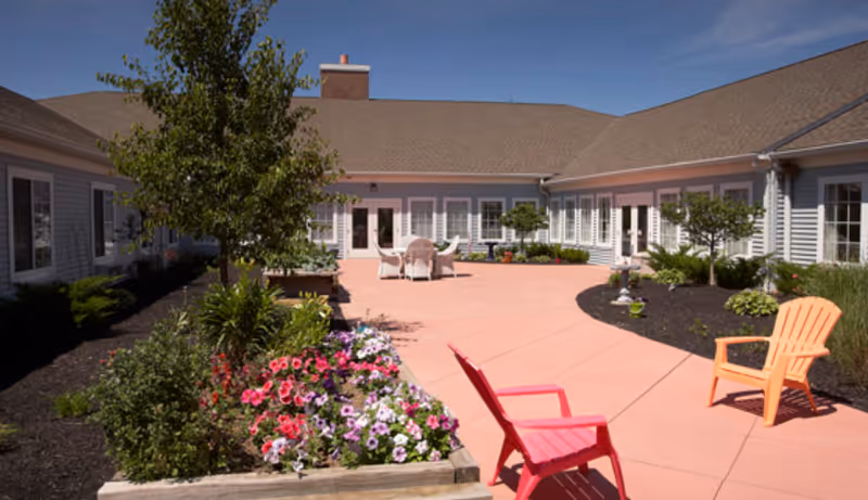 Outdoor courtyard area at a senior living facility with a paved patio, colorful flowers in raised garden beds, two plastic chairs in red and orange, and a table with chairs in the background. The building surrounds the courtyard with multiple windows and doors under a clear blue sky.