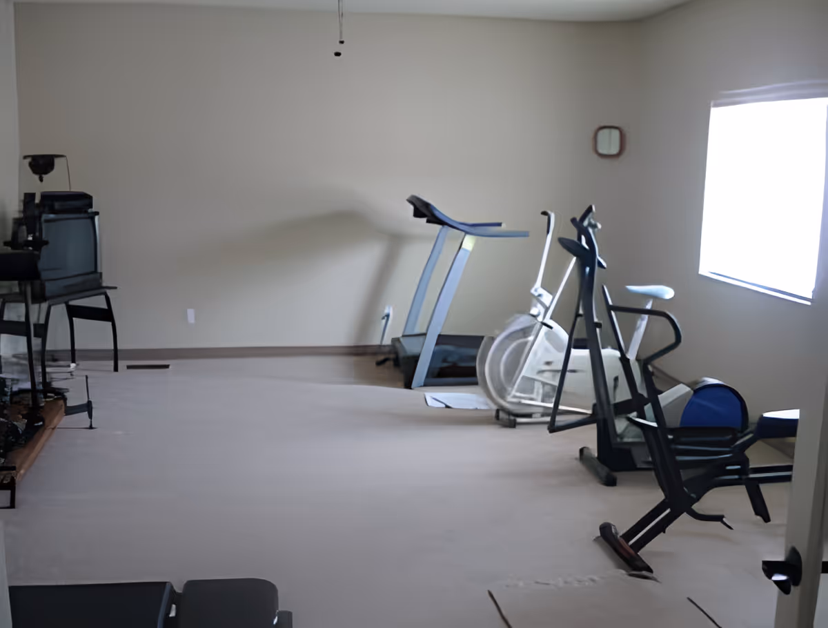 A small exercise room with a treadmill, stationary bike, and elliptical machine positioned near a window with natural light. The room has beige walls and carpeted floor, with a TV on a stand in the corner.