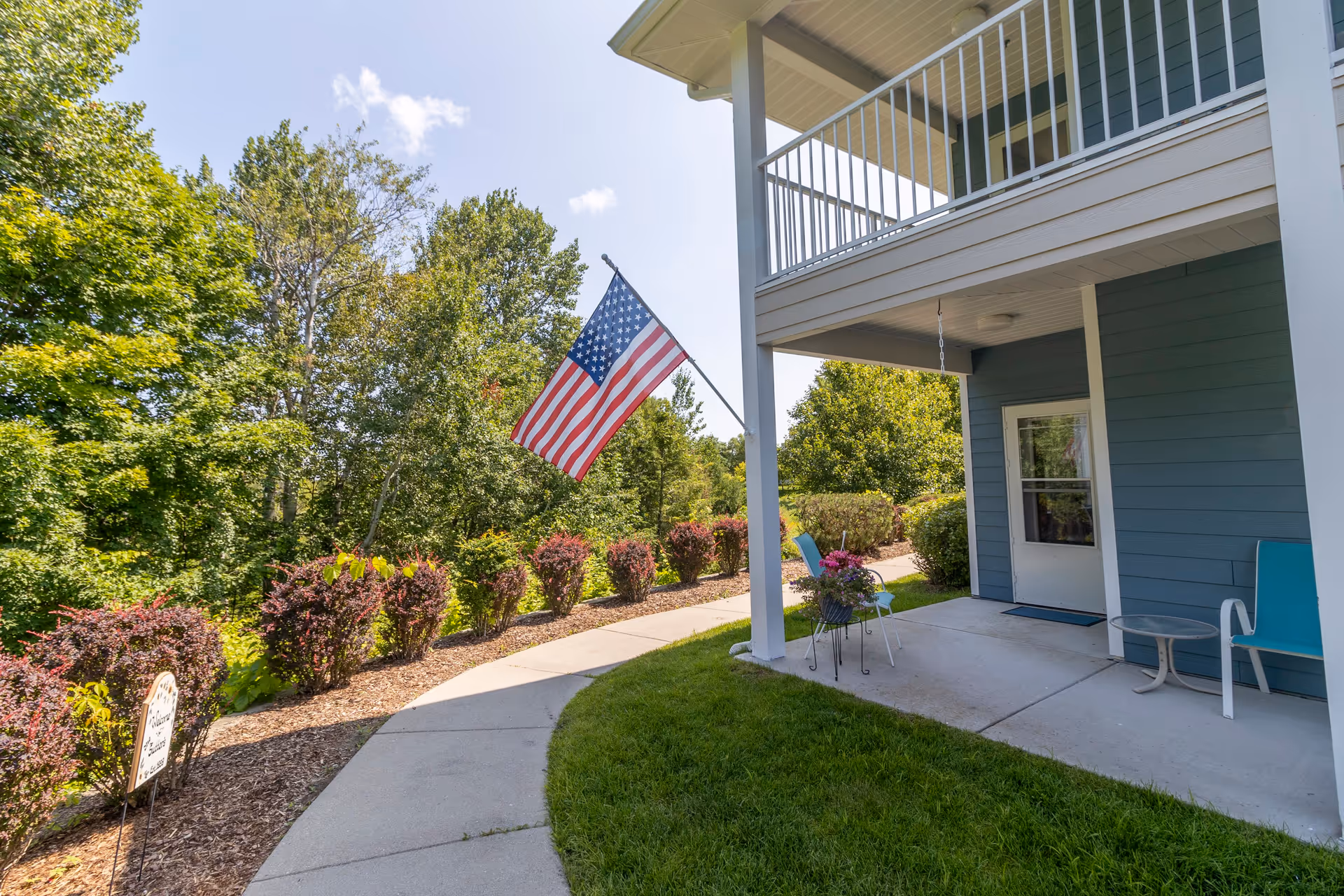 Outdoor patio area of a residential building with blue siding, a white door, and a balcony above. There are two chairs and a small table on the patio, along with a hanging flower pot. An American flag is mounted on a pole attached to the building, and a curved concrete walkway runs alongside landscaped bushes and trees under a clear blue sky.