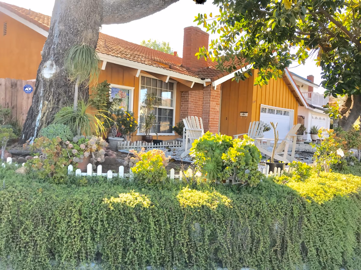 Front exterior view of a single-story house with an orange facade and a red tiled roof. There is a large tree in the front yard surrounded by various plants and shrubs. Two white wooden chairs are placed near the house entrance, and a white picket fence borders the garden area.