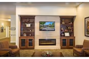 A cozy living room area featuring a wall-mounted flat-screen TV above a modern electric fireplace, flanked by built-in wooden shelves and cabinets displaying decorative items and framed photos. The room has warm lighting and comfortable seating with a brown armchair visible on the left.